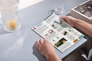 Close-up of a person reading an aesthetics conference newsletter at a clean desk, with a glass of water, lamp, and a stack of books nearby